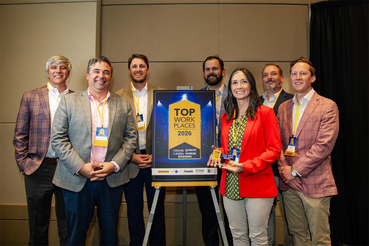 Team members from the firm’s Upstate and Coastal regions standing indoors beside a display reading ‘Top Workplaces 2026,’ with one member holding a matching award.