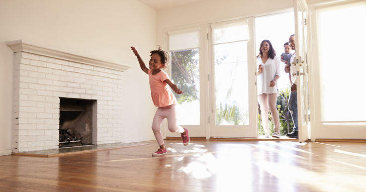 Child dances in empty living room beside fireplace while parents watch in doorway