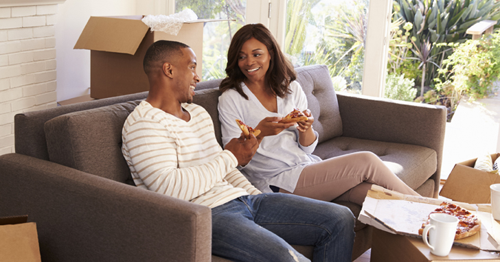 Couple eating pizza while sitting on sofa with moving boxes in background