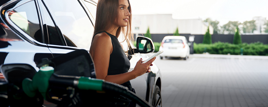 Young lady standing beside a car at a gas station, holding a phone near a fuel pump, with another vehicle and a building in the background.
