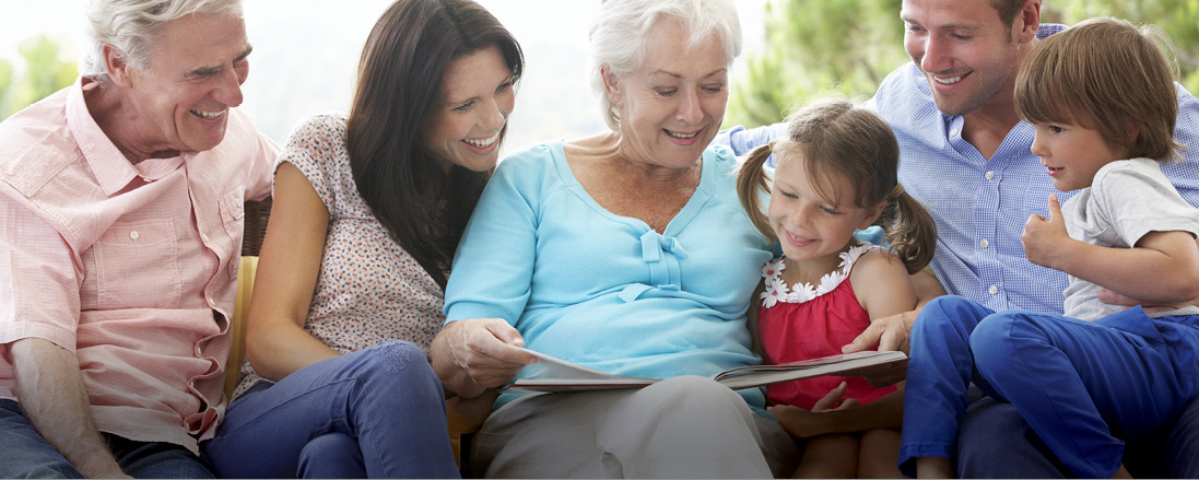 Multigenerational group seated together on a couch indoors, looking at an open book or album, with soft natural lighting and greenery visible in the background.