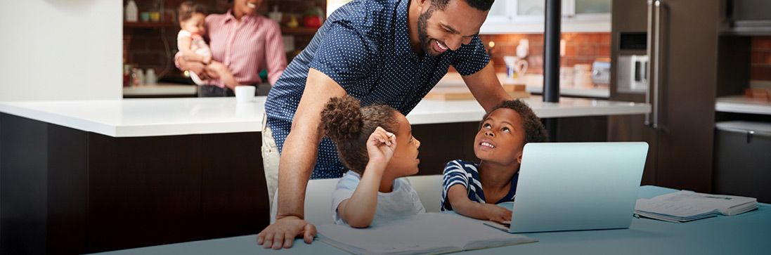 Adult assisting two children with homework at a kitchen table, with a laptop open in front of them; background shows another adult holding a baby.