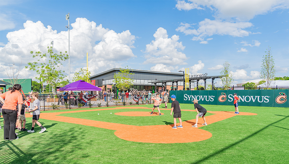 Children play a wiffle ball game on a small turf field inside a baseball stadium, with sponsor signage, spectators behind a fence, and a sunny sky overhead.